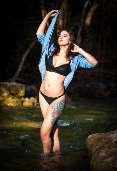 Standing in a shaded creek, black lace bralette under an open denim shirt, arms raised