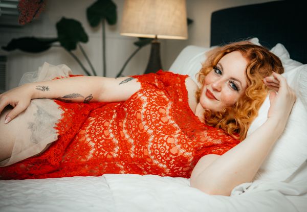 Lying on a white bed in a sheer orange lace dress, hair loose on the pillow