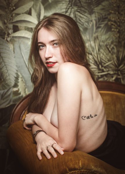 Seated in a vintage armchair, arms crossed over chest, botanical wallpaper backdrop