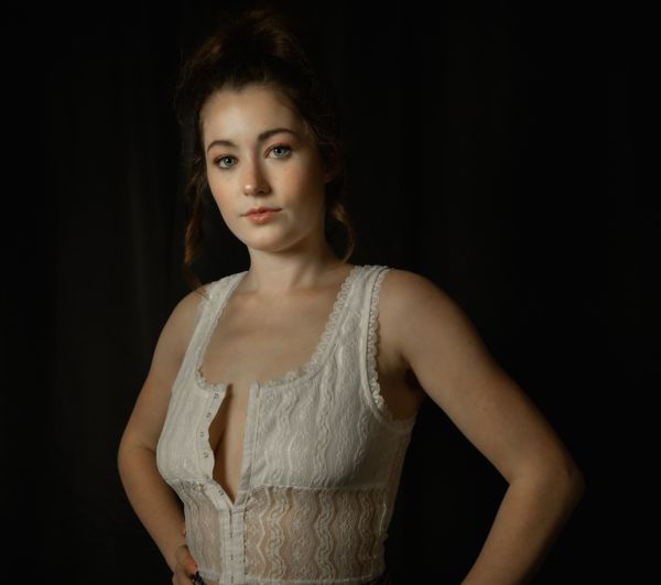 Studio portrait against black, hands on hips in a white lace corset top