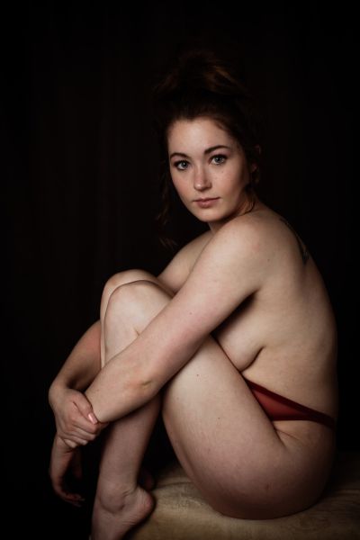 Seated profile portrait, knees drawn to chest, wearing a red lace thong, dark backdrop