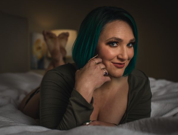 Lying on a bed in an open olive cardigan, chin resting on hand, soft lamp light
